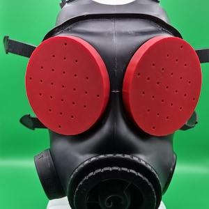 Könnte beinhalten: Eine schwarze Gasmaske mit zwei großen, roten, kreisförmigen Augenabdeckungen. Die Augenabdeckungen haben mehrere kleine Löcher. Die Maske hat verstellbare Riemen und eine kreisförmige Filterkomponente.