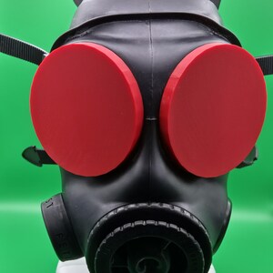 Könnte beinhalten: Eine schwarze Gasmaske mit großen, runden, roten Gläsern. Die Maske hat verstellbare Riemen und eine kreisförmige Filterkomponente. Die Maske besteht aus einem flexiblen Material und hat eine matte Oberfläche. Die Maske ist für den Schutz konzipiert.