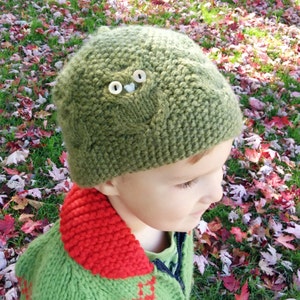 May include: A green knitted hat with a brown owl design and button eyes. The hat is worn by a child with a green and red sweater.