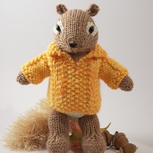 May include: A brown knitted squirrel wearing a yellow hooded sweater. The squirrel is standing on a brown leaf with acorns nearby.