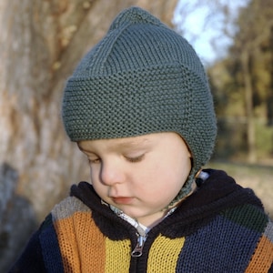 Peut inclure: Un jeune enfant portant un bonnet en tricot vert foncé avec des rabats d'oreilles et un pull rayé coloré.