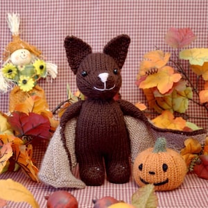 May include: A brown knitted bat with tan wings, a knitted orange pumpkin with a smiling face, and fall foliage.