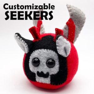 Può includere: Un giocattolo di peluche fatto a mano in rosso, nero e grigio, con grandi occhi neri e una faccia a forma di teschio. Il giocattolo ha piccole corna e ali, con il testo "Customizable SEEKERS" sopra.