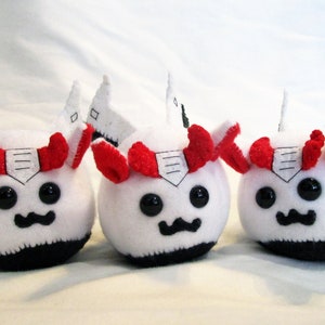 May include: Three plush toys with white bodies, black bottoms, and black eyes. Each has red horns, a white and black wing-like structure, and a black mustache. The toys are handmade.