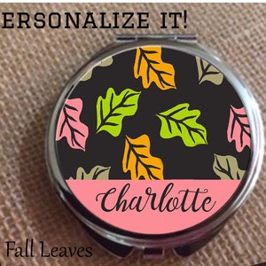 Peut inclure: Un miroir de poche avec un fond noir et un motif de feuilles d'automne colorées. La moitié inférieure du miroir est rose avec le nom "Charlotte" écrit en cursive noire.