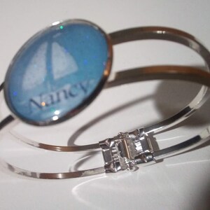 May include: A silver bangle bracelet with a blue glass charm featuring a sailboat and the name "Nancy" in white lettering.