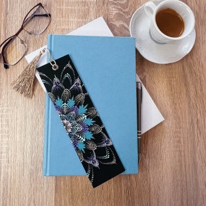 May include: A black bookmark with a colorful mandala design painted on it. The bookmark is lying on top of a blue notebook and a white piece of paper. A pen and a cup of coffee are also visible in the image.