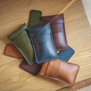 May include: Five leather phone cases in different colors, including green, blue, brown, and tan. The cases are arranged in a pile on a wooden surface.
