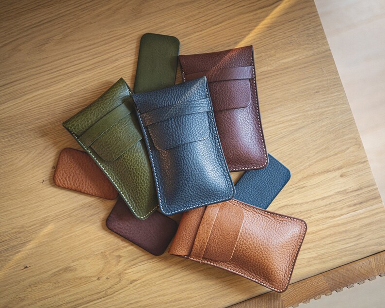 May include: Five leather phone cases in different colors, including green, blue, brown, and tan. The cases are arranged in a pile on a wooden surface.