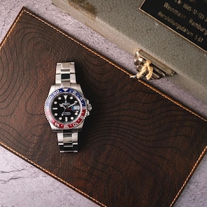 May include: A stainless steel wristwatch with a black dial and a red and blue rotating bezel, featuring a date window and a metal link bracelet, resting on a dark brown leather mat embossed with topographic lines next to a metal storage case.