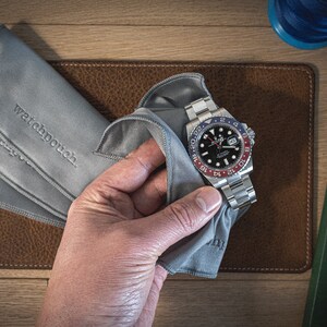 May include: A silver watch with a black face and a red and blue bezel. The watch is being cleaned with a gray microfiber cloth. The cloth has the words "watchpouch" embroidered on it.