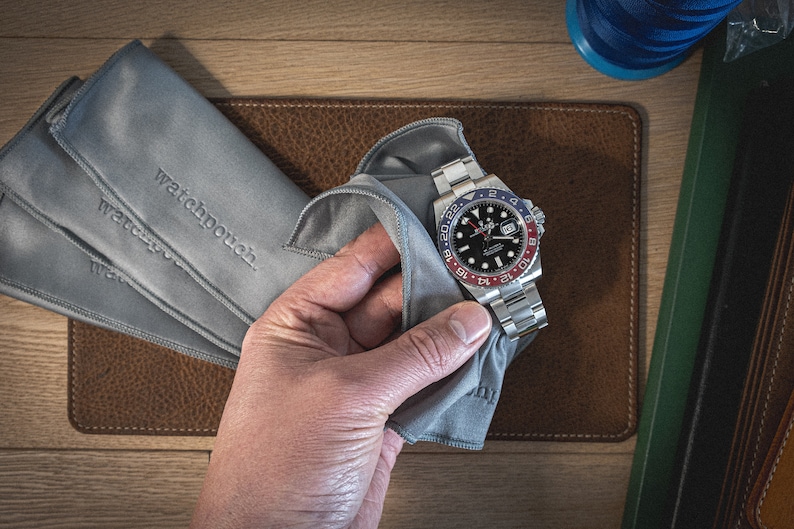 May include: A silver watch with a black face and a red and blue bezel. The watch is being cleaned with a gray microfiber cloth. The cloth has the words "watchpouch" embroidered on it.