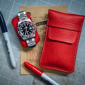 May include: A red leather watch case with a white stitch detail. The case is closed and sitting on a brown surface with a watch and a pen. The watch has a black face with blue and red accents.