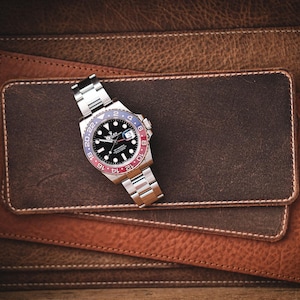 May include: A stainless steel wristwatch with a black dial and a red and blue bezel rests on a stack of brown leather wallets. The watch has a silver band and a date display. The wallets are various shades of brown.
