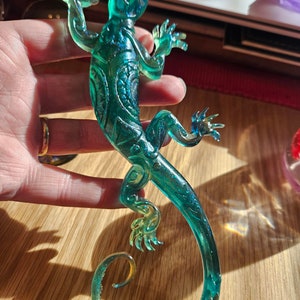 May include: A teal lizard ornament with detailed etched designs. The lizard's limbs are extended, and its tail is curled. The ornament is made of a clear material, allowing light to pass through, and is on a wooden surface.