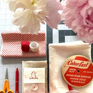 May include: A flat lay of sewing supplies including red and white gingham fabric, white fabric with a small embroidered house, a spool of white thread, a spool of red thread, a red pen, orange scissors, a red and white gingham ribbon, and a spool of white seam binding with the label "Colorfast 100 Yards Wash Fast Rayon Seam Binding Woven Edge Washable Made in U.S.A."