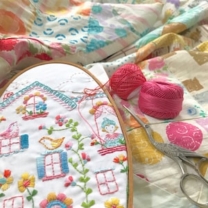 May include: A white embroidery hoop with a colorful embroidery design of a house with windows, birds, and flowers. Two pink balls of yarn and a pair of silver scissors are on a white fabric background.