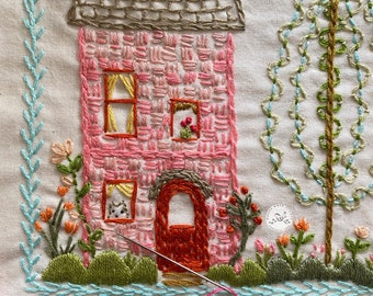 Puede incluir: Bordado que representa una casa rosa con un techo marrón, una puerta roja y un gato mirando por una ventana. La casa está rodeada de arbustos verdes y flores, y un árbol verde con hojas azules y verdes se encuentra a la derecha de la imagen.