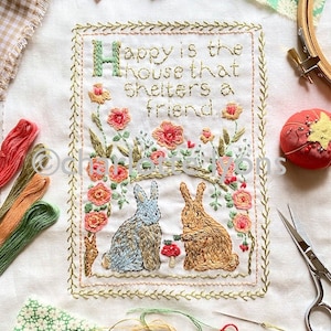 May include: Embroidered white fabric with a green frame and the text "Happy is the house that shelters a friend."  Two embroidered rabbits are sitting next to each other, one is blue and the other is brown.  There are pink and orange flowers embroidered around the rabbits.