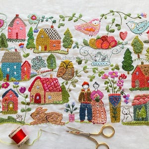 May include: Embroidered textile art featuring a village scene with colorful houses, trees, birds, and a rabbit. The artwork includes the text "charlotte lyons". A spool of red thread, needle, and scissors are also visible.