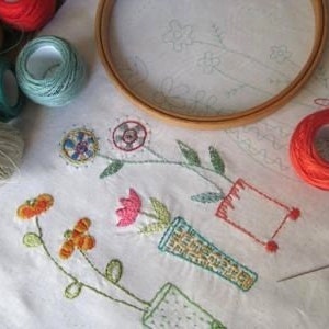 May include: A white fabric with embroidery of colorful flowers in vases. The fabric is surrounded by embroidery hoops, spools of thread, and needles.