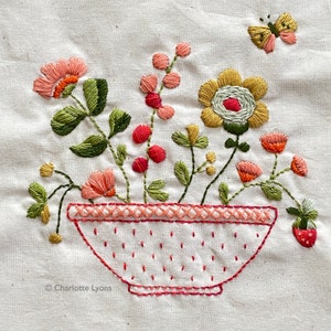 May include: Embroidered design of a bowl filled with pink and green flowers, a yellow flower, and a red strawberry. The bowl has a pink and white polka dot pattern and a pink and white border. The design is stitched on a white fabric background. The text "Charlotte Lyons" is in the lower left corner.
