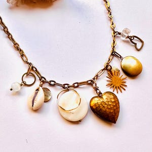 May include: A gold-toned chain necklace featuring various charms. Charms include a heart, sunburst, shell, and a round pendant. The necklace has a vintage aesthetic, with a mix of textures and shapes.