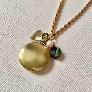 May include: A gold-toned locket necklace with a heart charm, a pearl, and a green abalone shell pendant. The round locket is closed and hangs from a delicate chain. The necklace is displayed on a white textured surface.
