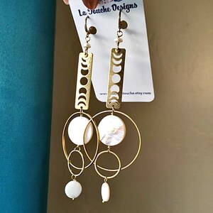 May include: Gold earrings with a moon phase design, white pearl accents, and a circle of white mother of pearl. The earrings are hanging from a card that says "La Touche Designs" and "latouche.etsy.com".