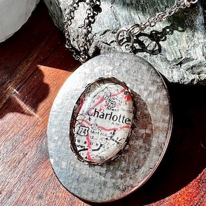 Puede incluir: Collar con colgante plateado con un mapa ovalado de Charlotte, Carolina del Norte. El mapa está encapsulado en resina transparente dentro de un marco plateado texturizado. El collar cuelga de una delicada cadena.