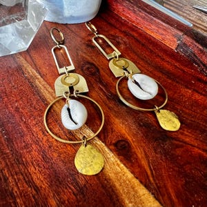 May include: A pair of brass earrings with a geometric design. Each earring features a rectangular top, a half-circle, a circular ring, and a teardrop-shaped charm. A white seashell is suspended within the circular ring. The earrings are displayed on a wooden surface.
