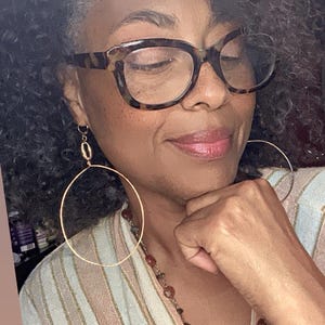 May include: Gold-tone hoop earrings. The earrings are large and circular, suspended from a smaller, decorative element. The person in the image is wearing tortoise shell glasses and a striped top with a necklace.