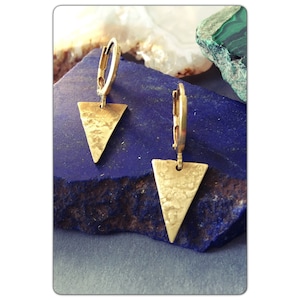 May include: A pair of gold hoop earrings with a hammered triangle pendant. The earrings are on a blue surface.