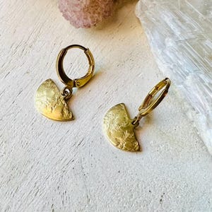 May include: A pair of gold hoop earrings with a textured, half-circle pendant. The earrings are made of metal and have a simple, modern design.