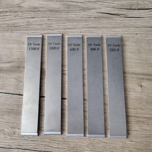 May include: Five rectangular metal sharpening stones with the text "SY Tools" and grit numbers 1500#, 1000#, 600#, 400#, and 220# printed on them. The stones are arranged vertically on a wooden surface.