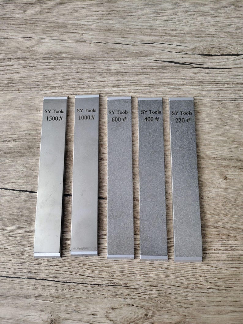 May include: Five rectangular metal sharpening stones with the text "SY Tools" and grit numbers 1500#, 1000#, 600#, 400#, and 220# printed on them. The stones are arranged vertically on a wooden surface.