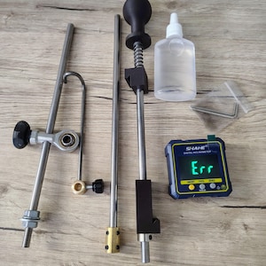 May include: An assortment of metal and plastic tools, including a digital inclinometer displaying "Err", a clear bottle, and various rods and connectors. The tools are arranged on a light wood-grain surface.