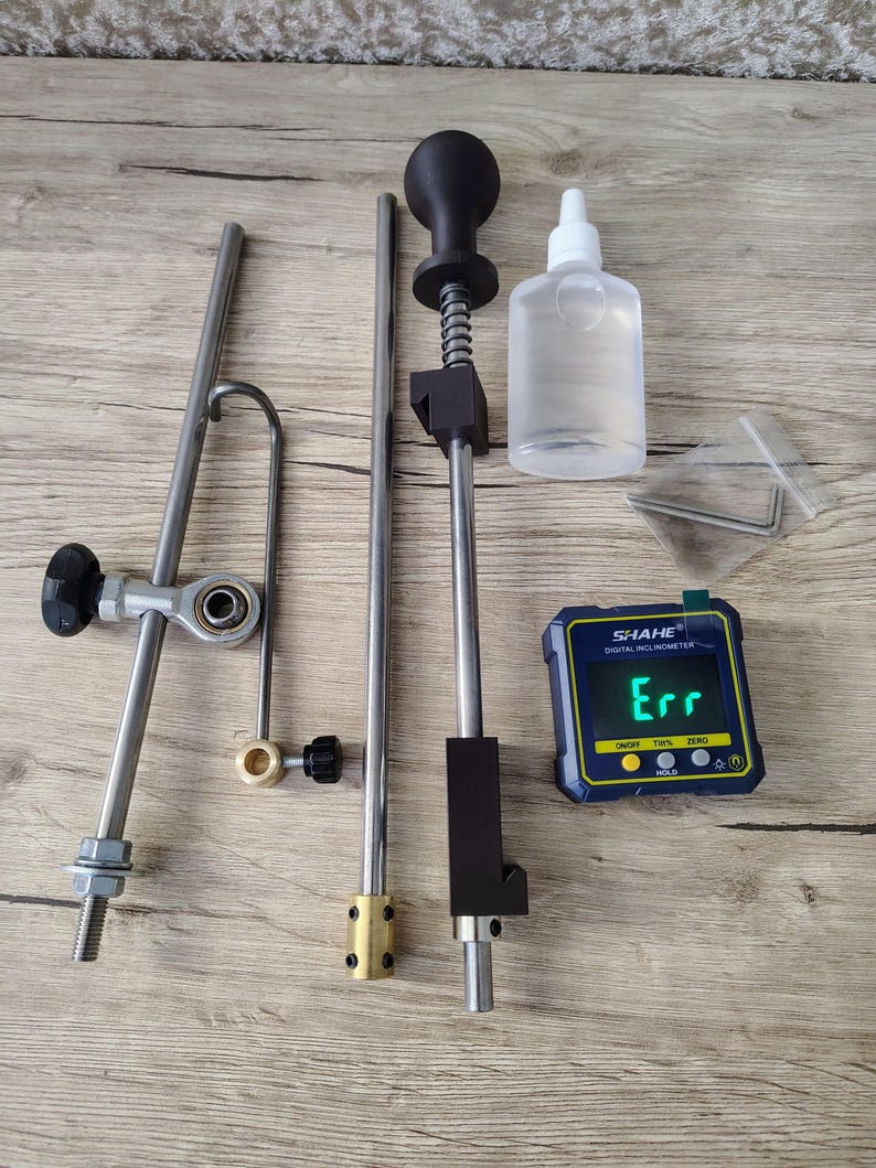 May include: An assortment of metal and plastic tools, including a digital inclinometer displaying "Err", a clear bottle, and various rods and connectors. The tools are arranged on a light wood-grain surface.
