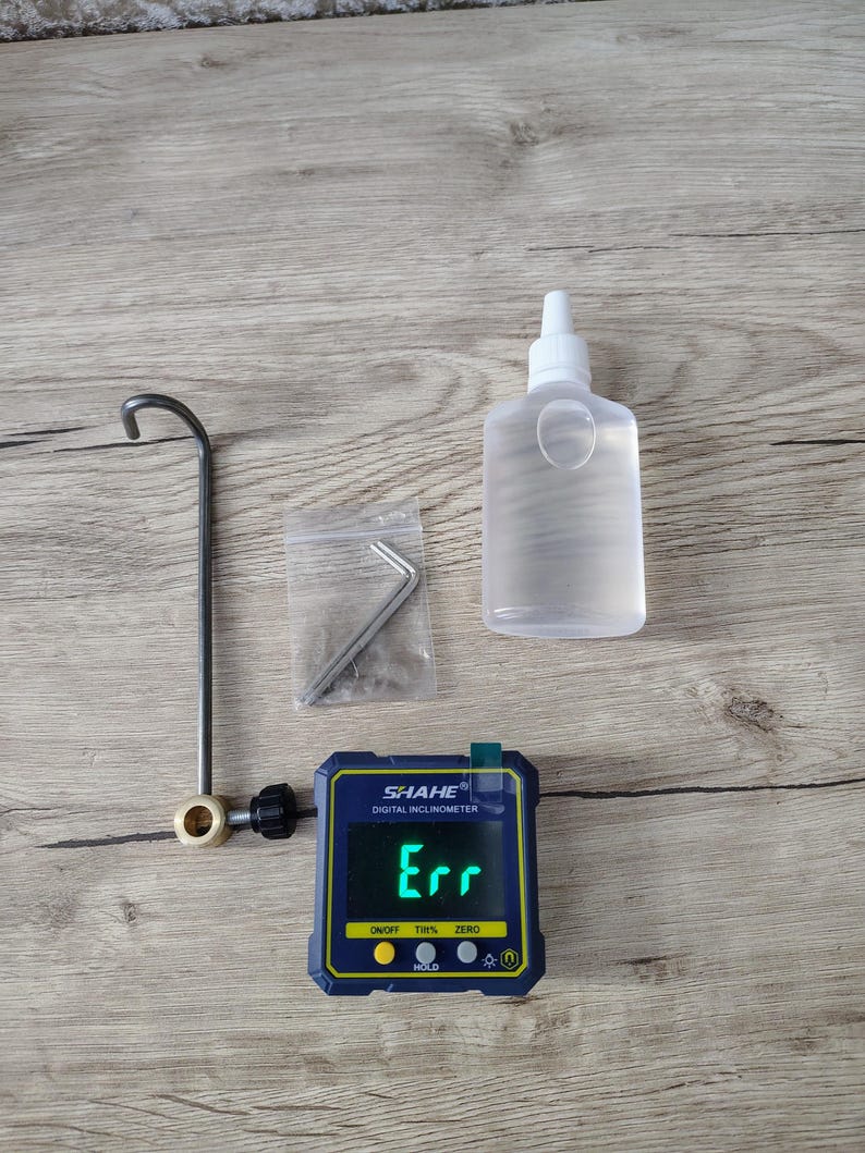 May include: A digital inclinometer with the text "Err" displayed on the screen, a clear plastic bottle, a metal tool, and a small bag with a metal tool. The inclinometer is blue and yellow.