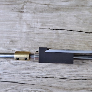 May include: A metal rod with a black, rectangular component and a brass fitting. The rod is positioned horizontally against a light brown wooden surface. The components appear to be part of a mechanical assembly.