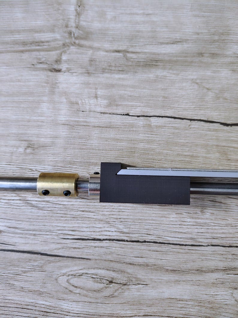 May include: A metal rod with a black, rectangular component and a brass fitting. The rod is positioned horizontally against a light brown wooden surface. The components appear to be part of a mechanical assembly.