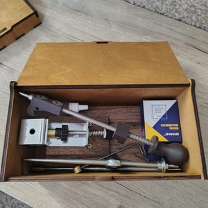 May include: A wooden box with a hinged lid, containing various metal and plastic tools. The tools include a silver-colored device with a black handle, a spring, and a digital inclinometer. The box is made of light brown wood.