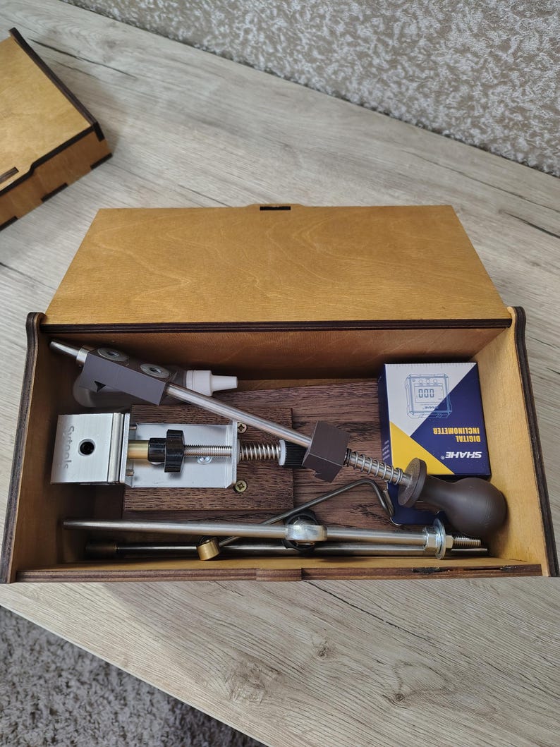 May include: A wooden box with a hinged lid, containing various metal and plastic tools. The tools include a silver-colored device with a black handle, a spring, and a digital inclinometer. The box is made of light brown wood.