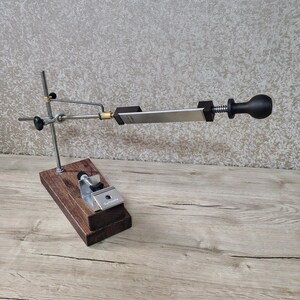 May include: A knife sharpening system with a wooden base, metal support arms, and a sharpening stone. The system includes a black handle and a silver-colored stone. The base is made of dark brown wood.