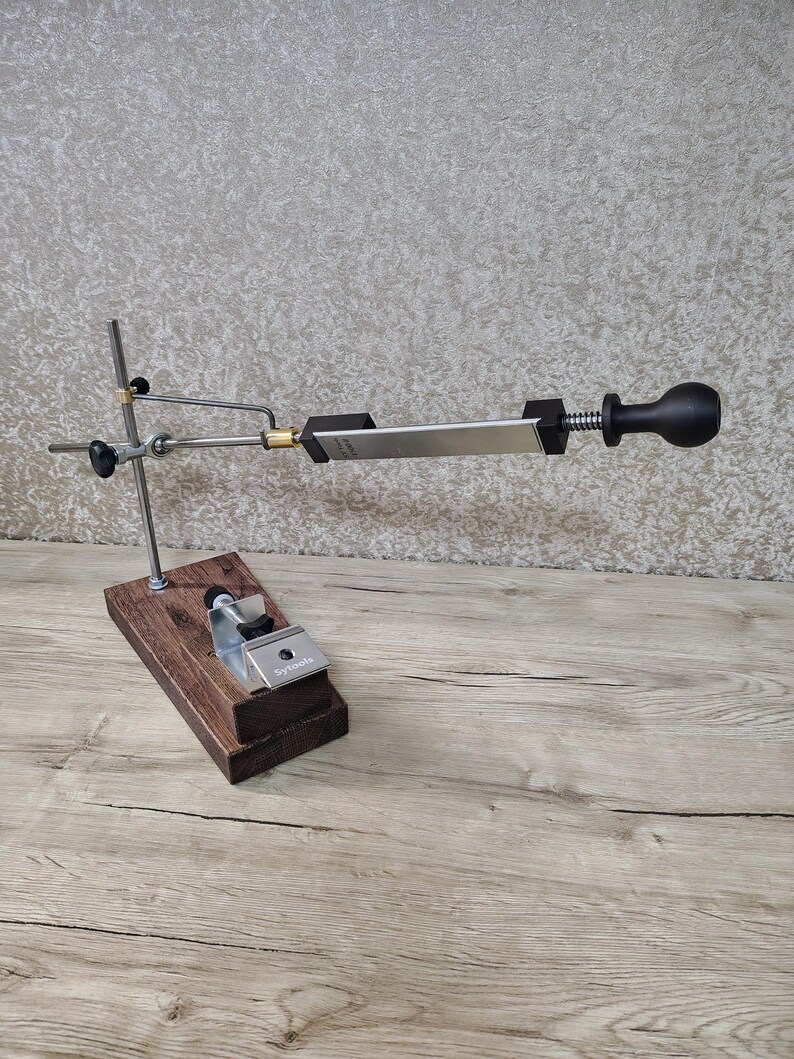 May include: A knife sharpening system with a wooden base, metal support arms, and a sharpening stone. The system includes a black handle and a silver-colored stone. The base is made of dark brown wood.