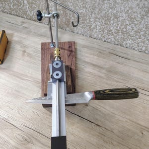 May include: A knife sharpening system with a stainless steel frame, wooden base, and a knife with a wooden handle. The system includes adjustable arms and a spring mechanism. The knife is positioned for sharpening.