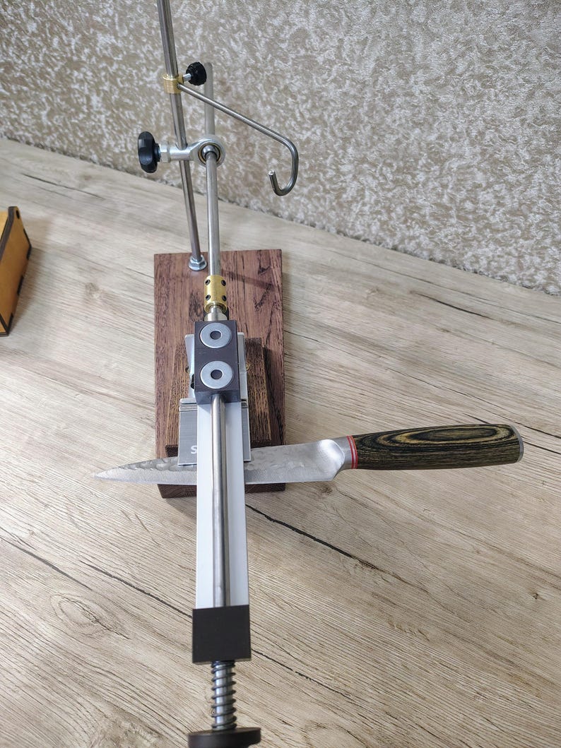 May include: A knife sharpening system with a stainless steel frame, wooden base, and a knife with a wooden handle. The system includes adjustable arms and a spring mechanism. The knife is positioned for sharpening.