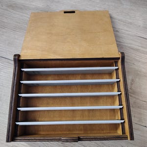 May include: A wooden box with a hinged lid, featuring a dark brown frame and a lighter wood interior. The box is divided into several horizontal compartments by thin, silver-colored dividers. The lid is open, revealing the interior.