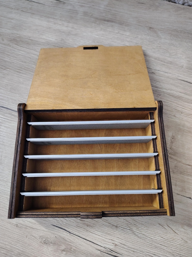 May include: A wooden box with a hinged lid, featuring a dark brown frame and a lighter wood interior. The box is divided into several horizontal compartments by thin, silver-colored dividers. The lid is open, revealing the interior.