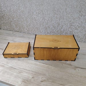 May include: Two wooden boxes with black trim. The larger box is rectangular, and the smaller box is square. Both boxes are made of light-colored wood and have a hinged lid. The boxes are sitting on a light-colored wooden surface.